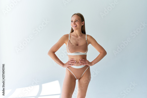Smiling adult woman in beige underwear standing with hands on hips in a bright minimalist room, presenting confidence, beauty and body positivity.