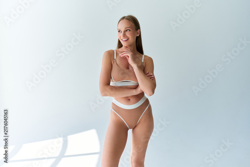 Smiling young woman in beige lingerie standing in a bright minimalist studio with soft sunlight and copy space.