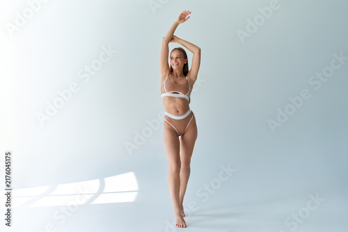 Adult woman in beige lingerie standing in a bright minimalist studio, stretching with raised arms as sunlight casts window shadows across the floor and wall.