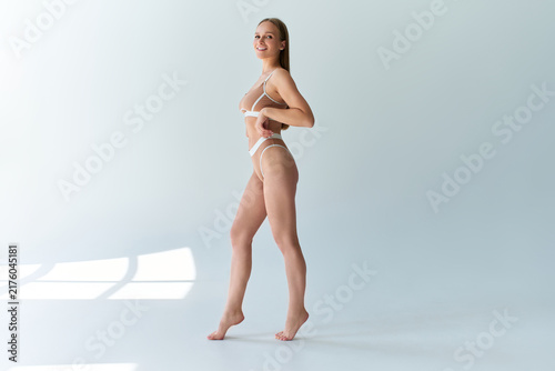Smiling adult woman in beige lingerie standing in a bright minimalist studio, with soft sunlight and window shadows creating a clean fashion and beauty setting.