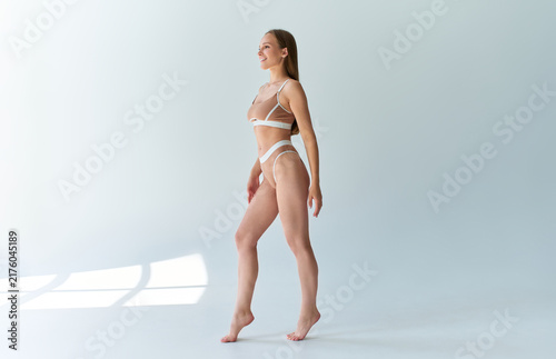 A smiling, beautiful young woman in comfortable beige underwear poses barefoot in a minimalist studio with soft window shadows and plenty of open space.