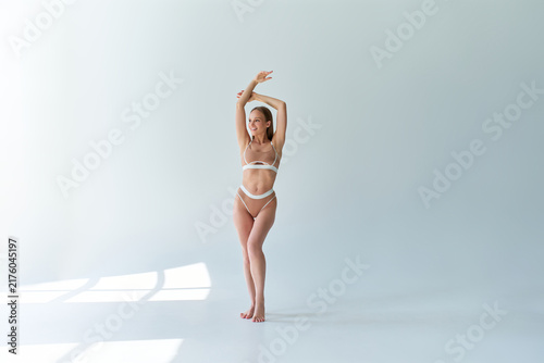 Woman in beige swimwear posing with raised arms in a bright minimalist studio, with soft window shadows across the floor and wall.