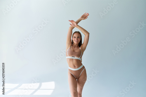 Young woman in beige lingerie standing with arms raised in a bright minimalist room, expressing confidence, wellness, and body positivity.