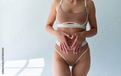 Cropped woman in beige lingerie standing indoors with hands forming a heart over her abdomen, lit by soft sunlight and window shadows.