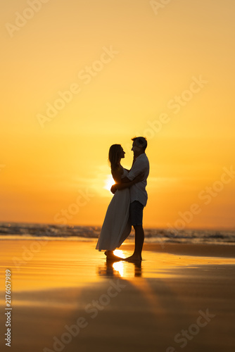 Couple stands on the beach at sunset with waves in the background and warm colors in the sky