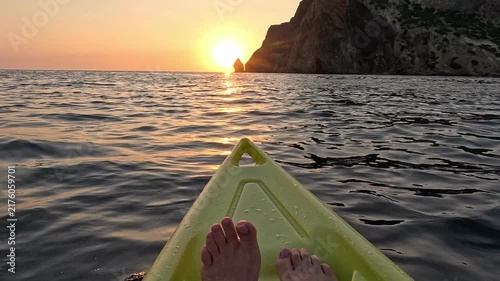 Sailing yellow kayak past rocky coastline during vibrant sunset. First-person POV wide shot.