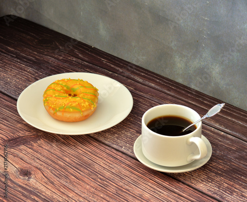 Canvastavla A fresh doughnut with yellow linoleum filling and topping and a cup of hot black coffee on a wooden table