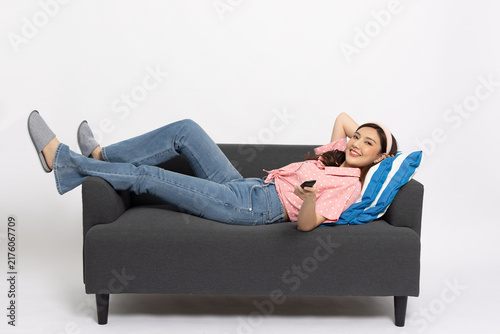 Happy young Asian woman lying on couch holding remote control, watching television show and relaxing at home isolated on white background, enjoying comfortable lifestyle and weekend leisure time