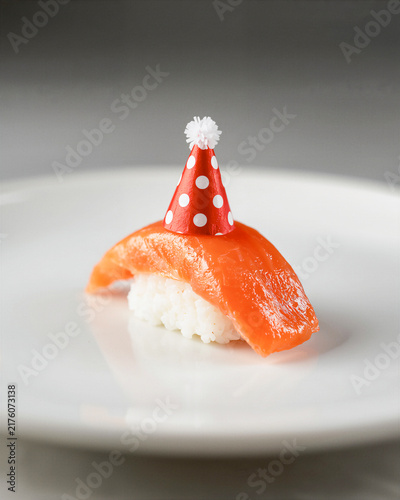 Sushi on white plate background. Salmon nigiri wearing Happy Birthday hat. Christmas, New Year celebration party. Creative Japanese food miniature. Red polka dot cap. Vertical, space for design