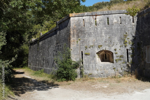 Photography A massive stone wall of Fort Liédot featuring narrow embrasures and embrasure slits stands surrounded by green foliage along a dirt path under direct sunlight