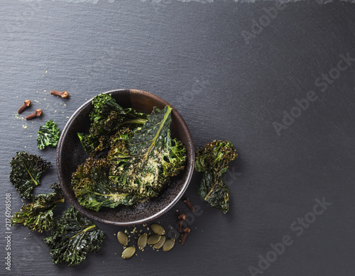 Delicious homemade crispy kale chips served in a rustic bowl accompanied by pumpkin seeds and aromatic cloves on a dark natural slate background for a healthy nutritious snack