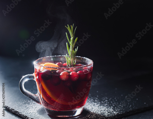 Warm and inviting steaming cranberry beverage garnished with fresh orange slices and aromatic rosemary in a transparent glass mug on a sleek dark surface