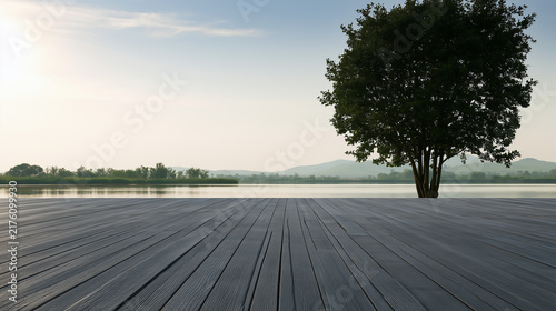A dark gray wooden boardwalk in the foreground leads to a calm lake with a solitary green tree on the right and distant mountains.