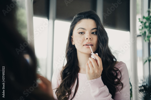 Fotografía Young woman applying lip gloss in front of mirror at home, getting ready with na