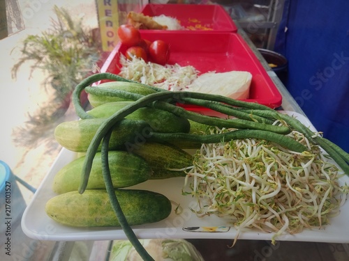 A close up of various green vegetables fresh cucumber, yardlong beans, and raw bean sprout arranged in a plastic container for cooking