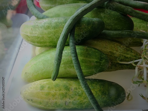 A close up of various green vegetables fresh cucumber, yardlong beans, and raw bean sprout arranged in a plastic container for cooking