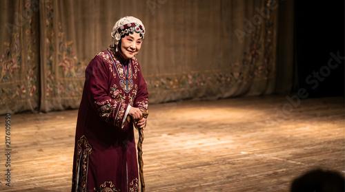 An elderly female opera performer in traditional embroidered costume stands on a wooden stage with a walking stick, wearing stage makeup and headdress.