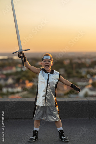 Canvas Print Excited young boy in futuristic silver costume holding a medieval sword aloft on