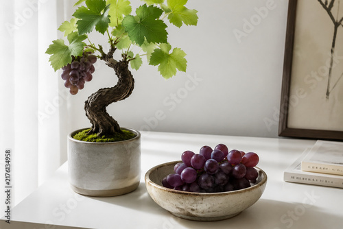 Potted grapevine with curved trunk and ceramic bowl of purple grapes on white desk, home gardening and harvest concept.