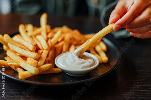 Close-up of a hand dipping a golden French fry into creamy seasoned sauce beside a plate piled with crispy fries at a restaurant table