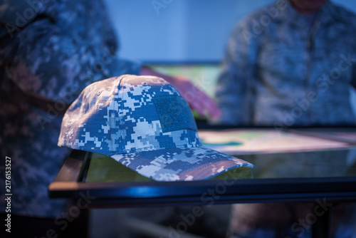Digital camouflage military cap on a table during a briefing, with uniformed service members and documents blurred in the background.