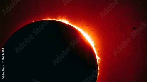 A close-up view of a total solar eclipse, revealing the Sun's fiery corona and prominences glowing intensely around the dark silhouette of the Moon against a deep red sky.