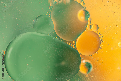 Abstract macro photograph of oil bubbles floating in liquid, creating large translucent green and yellow circles with tiny droplets and a vibrant colorful background.