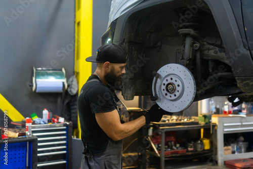 Lifted car reveals brake rotor and suspension inside workshop bay. Bearded mechanic tightens caliper bolts checking wheel hub with wrench
