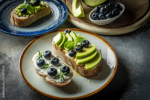 Avocado and blueberry toast on rustic sourdough bread served for healthy breakfast. Gourmet plant-based open sandwich with mint leaves decoration on a plate, top view food photography.