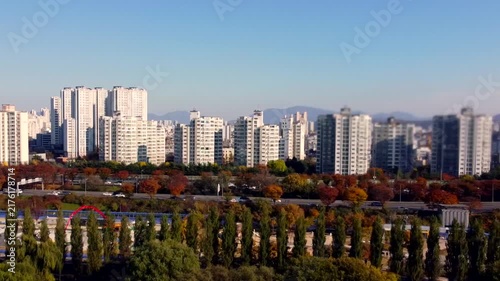 Modern residential skyline borders autumn park, with high-rise apartment towers rising behind colorful trees. Dense urban housing contrasts fall foliage, creating scenic city and nature view.