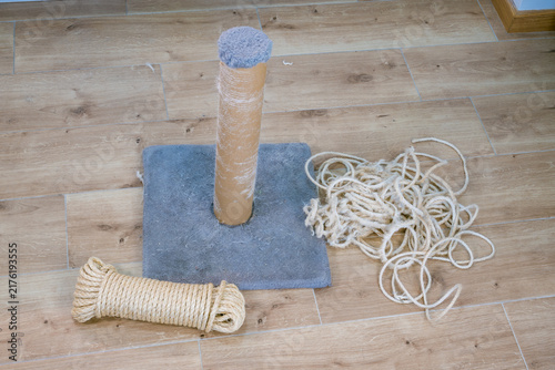 Disassembled cat scratching post with cardboard cylinder, blue carpet base, loose natural-fiber rope, and replacement cord on an indoor wooden floor.