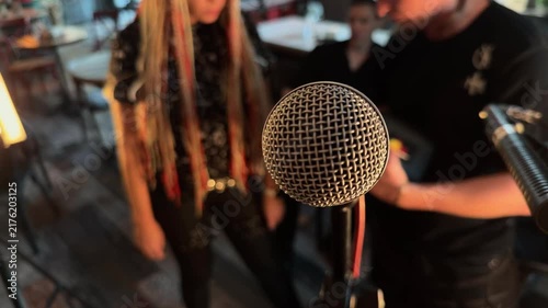 Professional vocal microphone close up on stand during live band soundcheck with blurred unrecognizable musicians preparing in background inside cozy music venue handheld orbiting documentary arc shot
