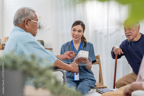 Asian female caregiver measuring blood pressure for senior wheelchair patient during health checkup in retirement home. Elderly healthcare routine checkup with professional nurse
