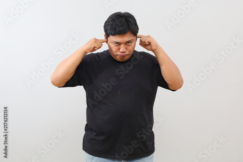Obraz na plátne A heavy-set man with a sad expression covers his ears with his fingers to avoid hearing anything, shown against a plain white background