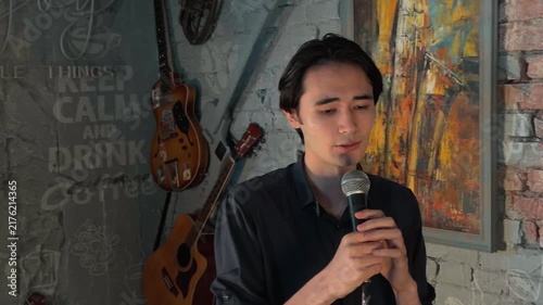 Passionate young Asian male spiritual leader preaching motivational sermon from a stage in a cozy intimate cafe club environment, holding a wired microphone with unstable handheld camera movement