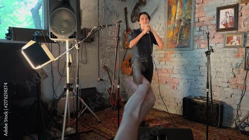 Expressive Asian Male Singer Performing Live Soundcheck On Cozy Indie Cafe Stage While Unrecognizable Drummer Spins Drumstick In Foreground During Unplugged Acoustic Jazz Gig Authentic Handheld Shot
