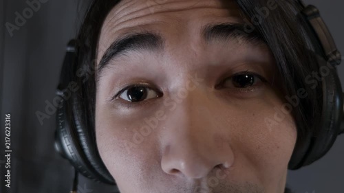 Intense young Asian male podcast host wearing black headphones speaking emotionally into the camera recording true crime audio episode inside dark modern digital media studio extreme close up shot