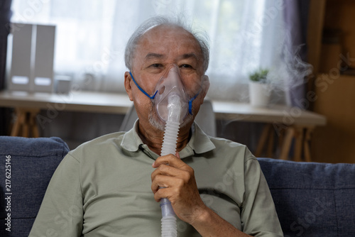 Elderly Asian man using nebulizer mask at home, receiving respiratory treatment while seated on sofa with calm expression and visible vapor in domestic setting