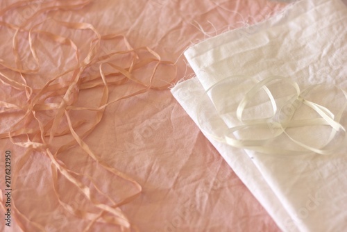 Avocado-dyed cotton fabric serves as the background, displaying a soft pink hue. On top, there is a neatly folded piece of white cotton fabric. Thin silk ribbons are scattered across, adding texture