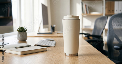 A modern office workspace featuring a disposable coffee cup on a wooden desk with an open notepad, keyboard, and a small plant in the background.