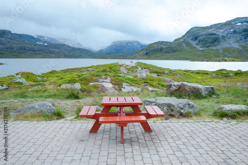 Quadro su tela Red wooden picnic table on a paved area overlooking a vast mountain lake and rocky hills under a cloudy summer sky in a rural Scandinavian landscape