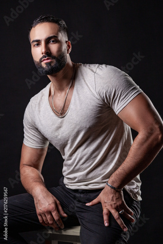 Confident guy in t-shirt sitting with hands on knees showing strong muscular arms wearing silver chain and bracelet in cinematic lighting isolated over black background