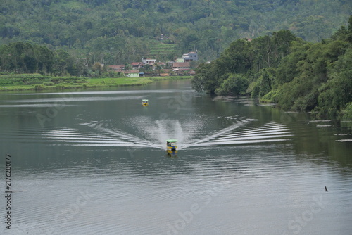A tranquil tropical lake surrounded by dense lush rainforest and green hills, with a small traditional boat cruising across calm water.