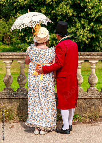 A couple dressed in Regency-era historical costumes stands together, enjoying a stroll in a garden, reflecting the elegance and romantic atmosphere of the 19th century.