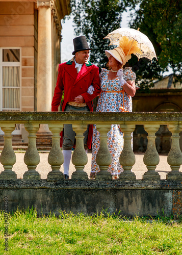 Fotografía A man and woman dressed in 19th-century Regency-era attire walk arm-in-arm through an estate garden, likely participating in a historical reenactment or themed cultural event