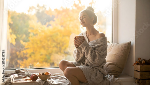 Thanksgiving & Autumn Wellness: Peaceful woman relaxes by window with warm beverage, enjoying golden autumn view