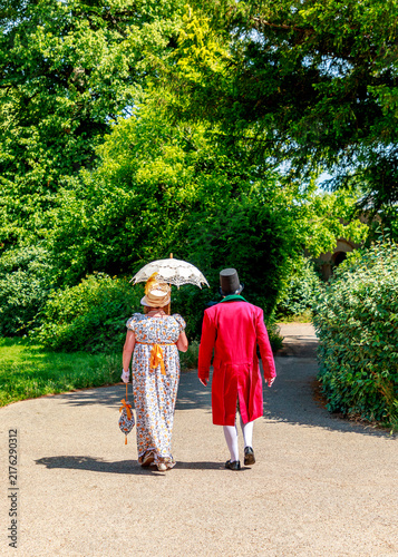 Картината върху платно A couple dressed in elegant 19th-century period costumes strolls down a scenic park path, likely participating in a historical reenactment or formal themed event