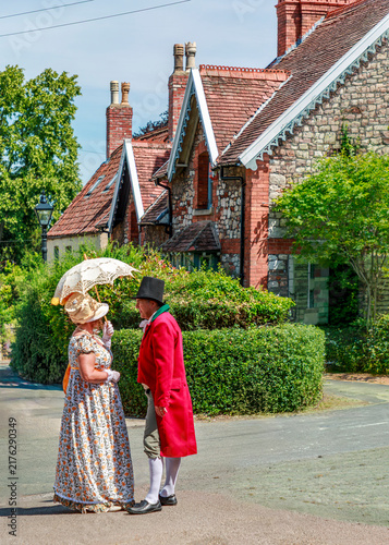 Photography A couple in 19th-century attire, including a red frock coat and a lace parasol, converse in front of historic houses, likely attending a historical reenactment event