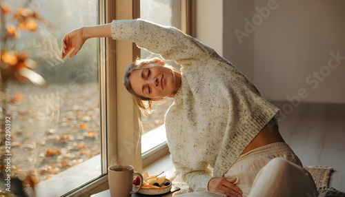 Thanksgiving & Autumn Wellness: Young woman stretching peacefully near window. Enjoying autumn wellness, embracing calm and joy