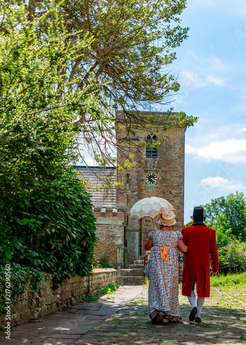 Fotografie A couple dressed in authentic Regency-era attire walk arm-in-arm toward an old stone church, likely participating in a historical reenactment or themed event in the English countryside